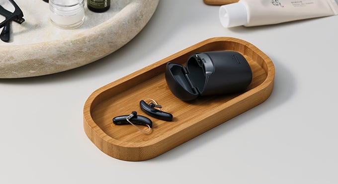 A pair of sleek black hearing aids with a charging case sit on a wooden tray beside grooming items and eyewear on a light-colored surface.