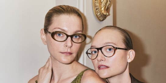 Two women with slicked-back hair wearing Christopher Esber glasses stand closely together indoors, facing the camera.