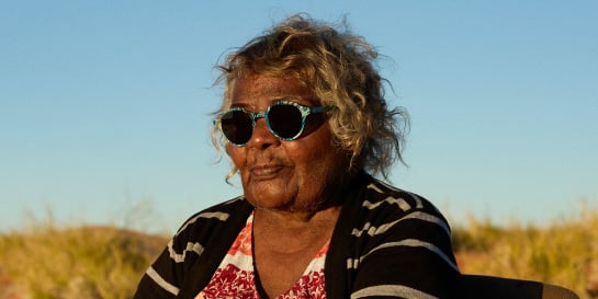 Aboriginal artist Corban Clause Williams wearing sunglasses in front of a colorful, abstract background with circular patterns.