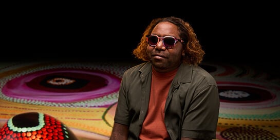 Aboriginal artist Corban Clause Williams wearing sunglasses in front of a colorful, abstract background with circular patterns.