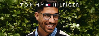 Man with dark hair and beard wearing blue glasses smiles in front of green leafy background with 'Tommy Hilfiger' logo above.