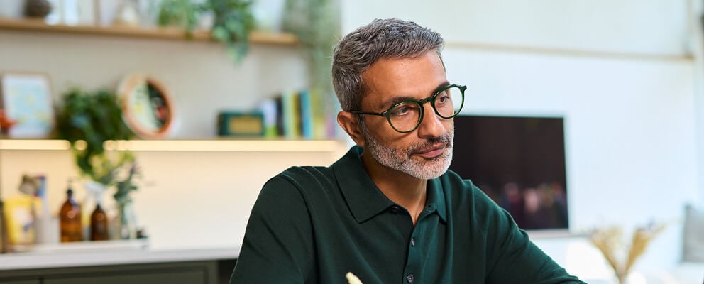 A man with gray hair and glasses sits at a desk, writing in a notebook with a pen. A laptop and shelves with books and plants are in the background.
